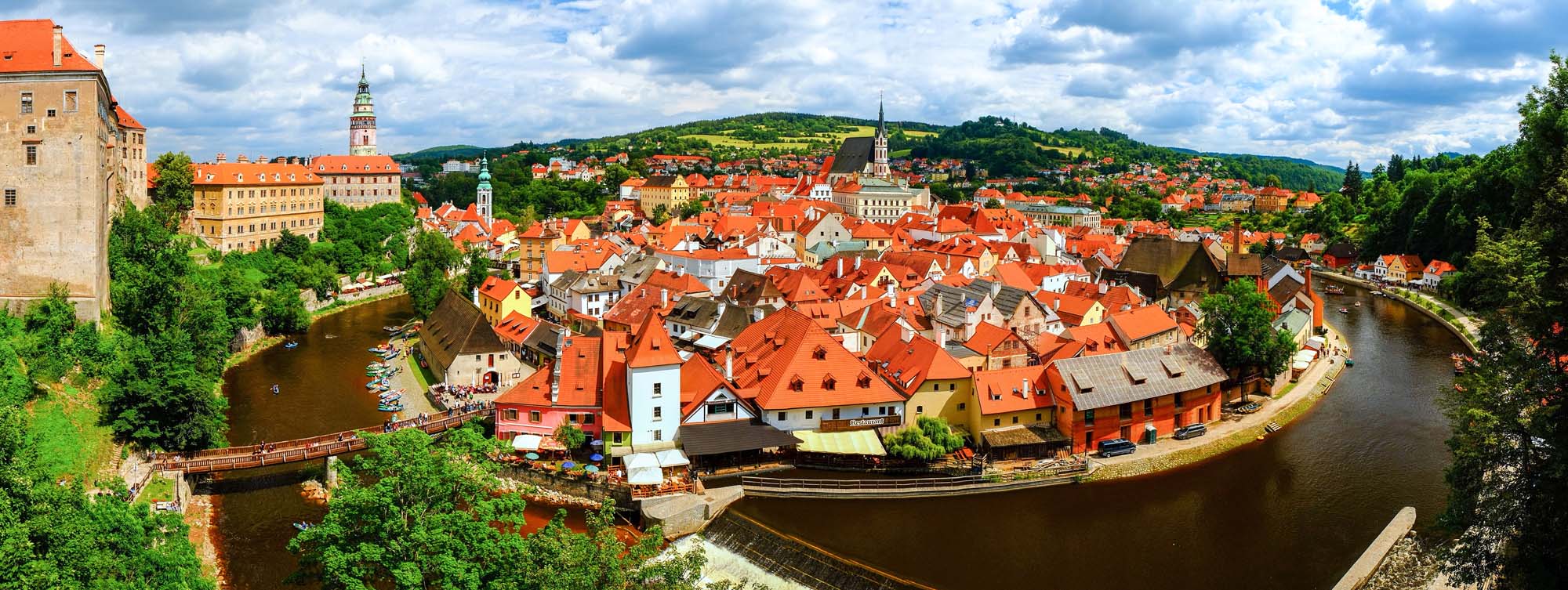 A stunning panoramic view of Cesky Krumlov, a UNESCO World Heritage Site in the Czech Republic, featuring the iconic red-tiled roofs of the medieval old town nestled within a dramatic bend of the Vltava River, with the historic castle and church spires rising prominently against forested hills under a partly cloudy sky.