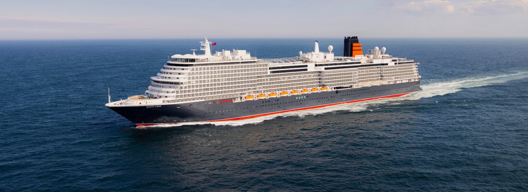 queen-anne-at-sea-2880×1047-desktopjpgimage2340851high Queen Anne Cruise Ship at Sea - A large modern cruise ship, the Queen Anne, sails through open ocean waters with a distinctive white superstructure and dark hull accented by a bold red stripe, characteristic of Cunard Line's fleet design.