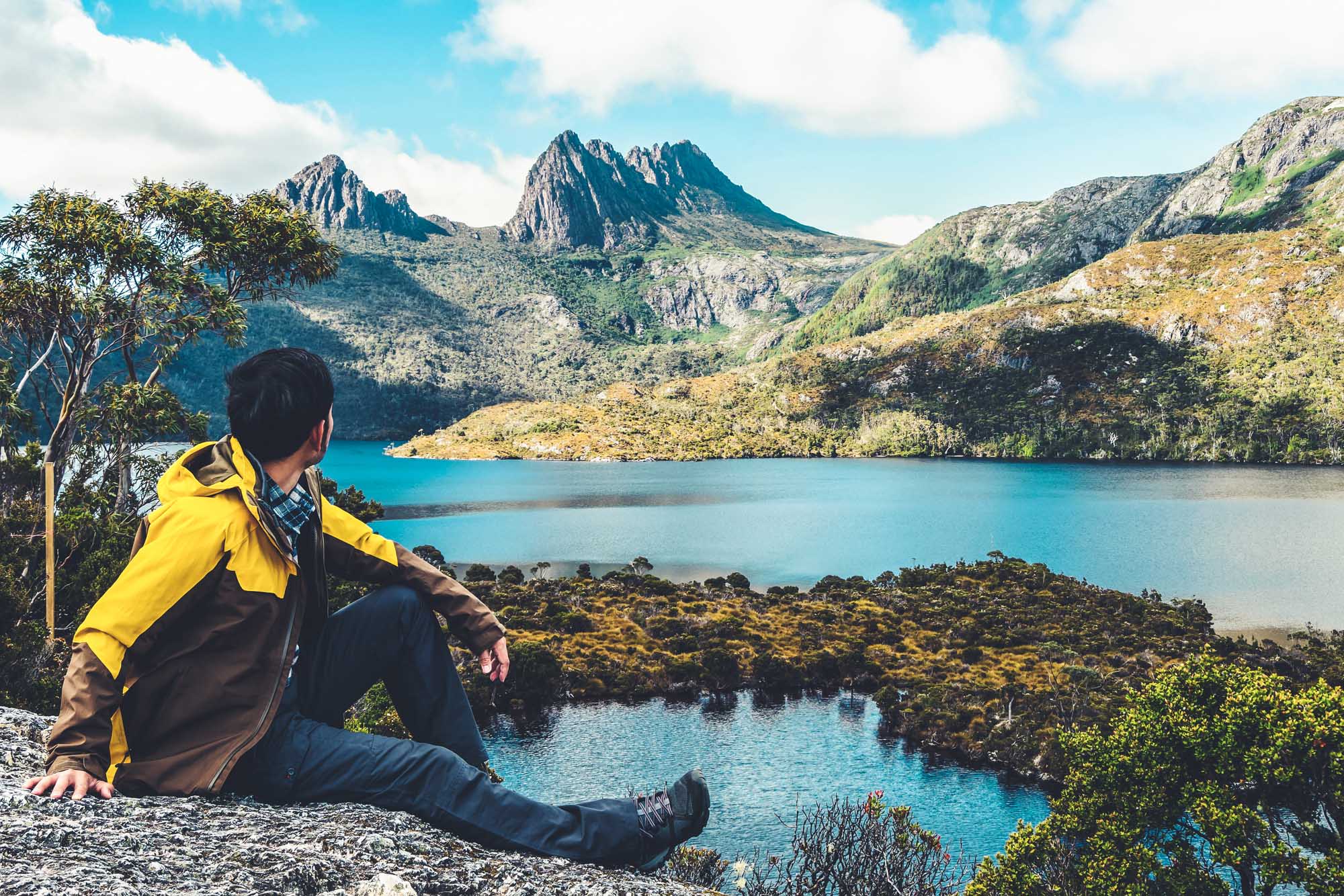 A solo traveler in a bright yellow jacket sits on a rocky outcrop overlooking the stunning
