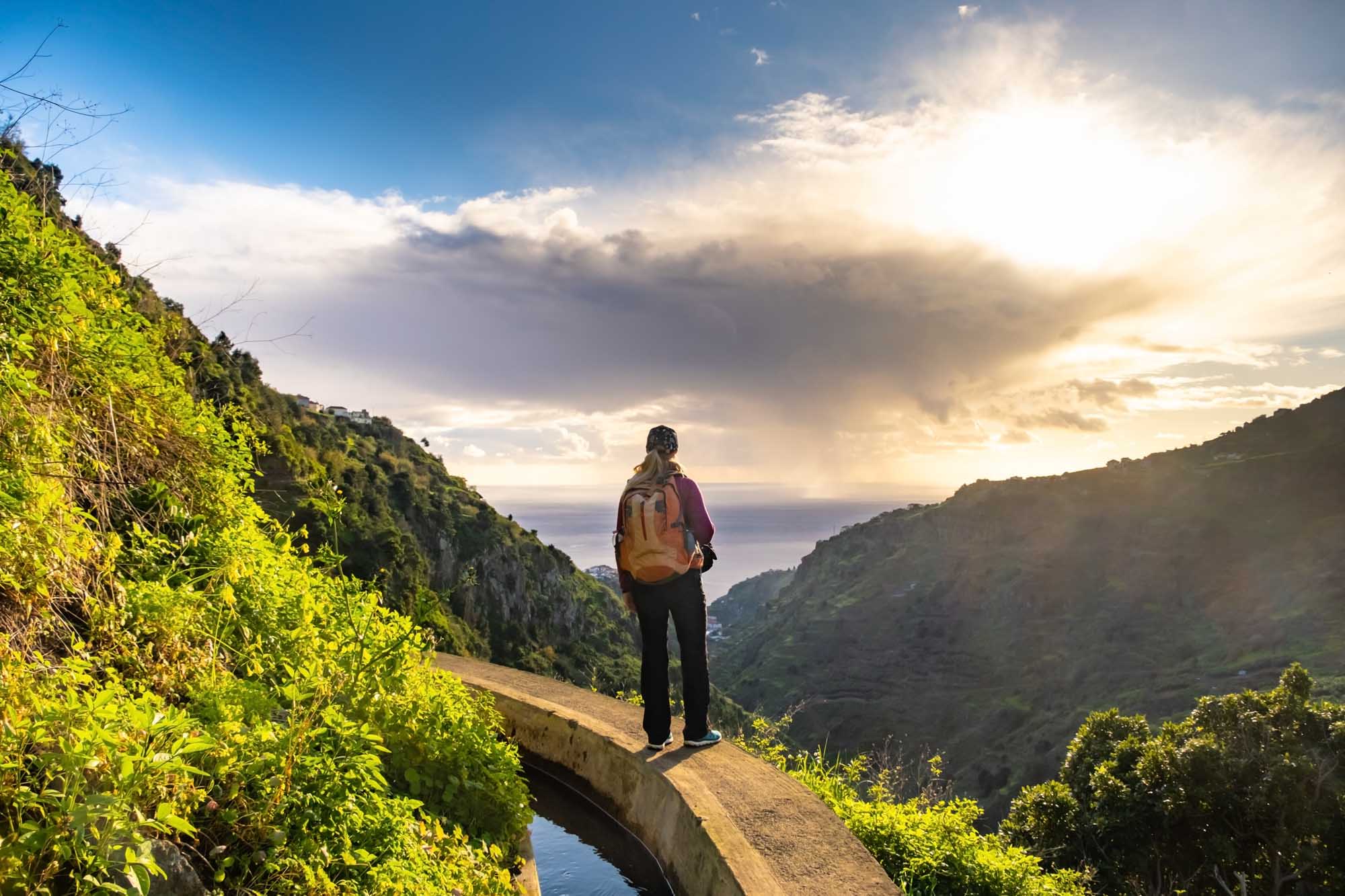 A solo hiker with an orange backpack stands on a narrow concrete path alongside the Levada do Norte