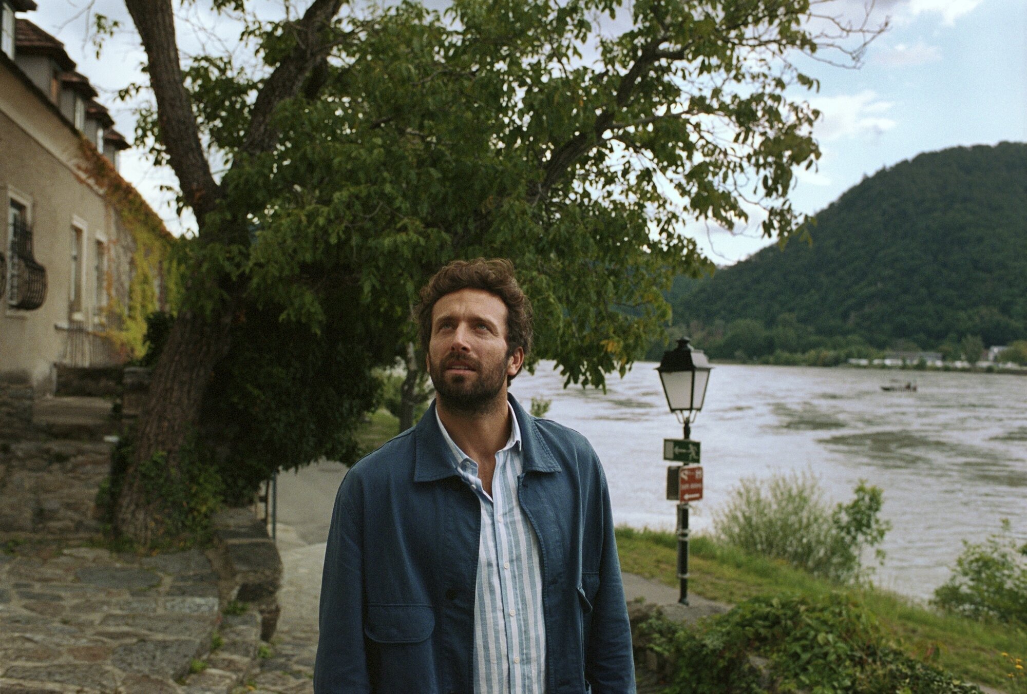 A bearded man in casual attire stands along a picturesque riverside promenade beneath weeping